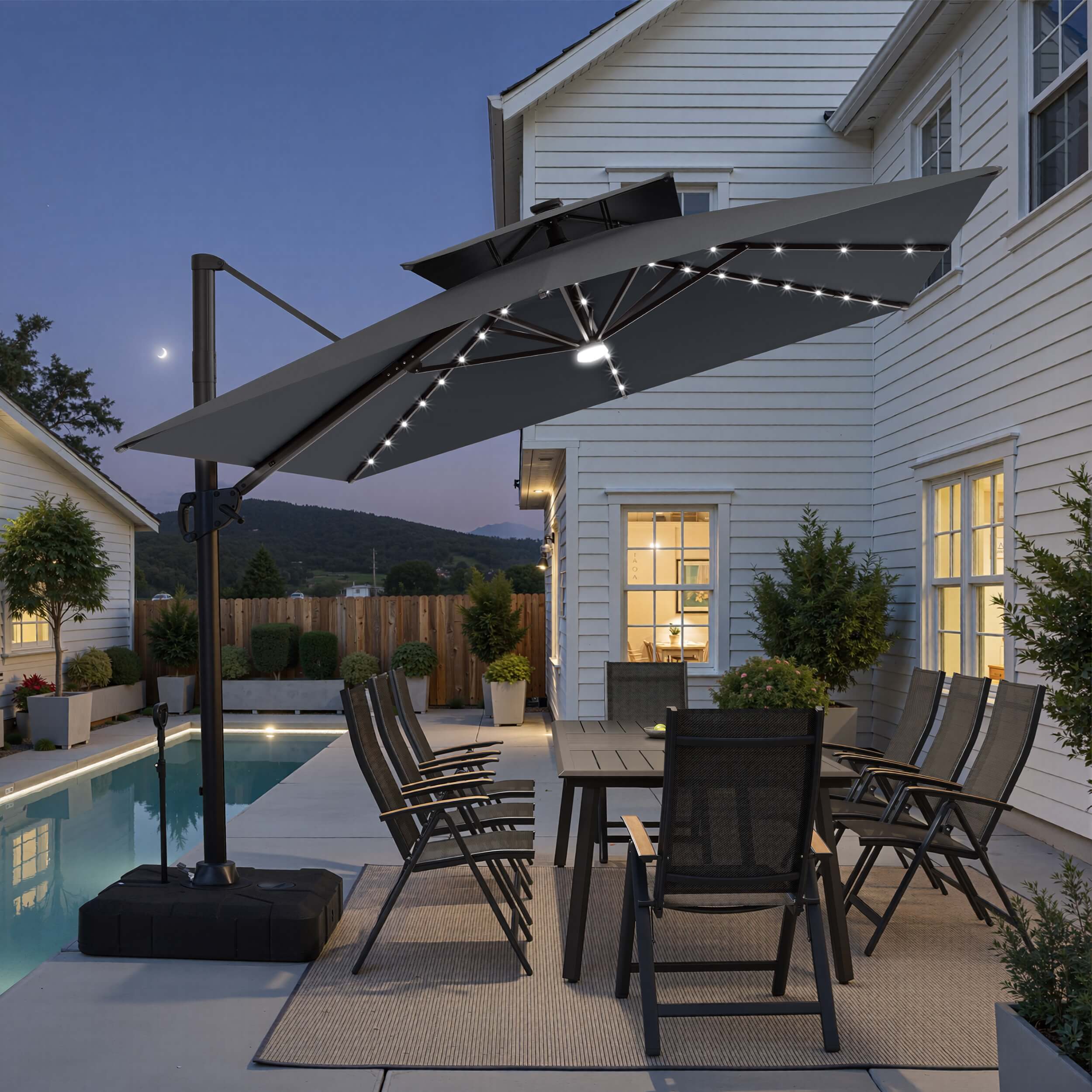 Tilted 12x9 ft grey lighted cantilever umbrella with wheeled base shading a poolside dining set at twilight